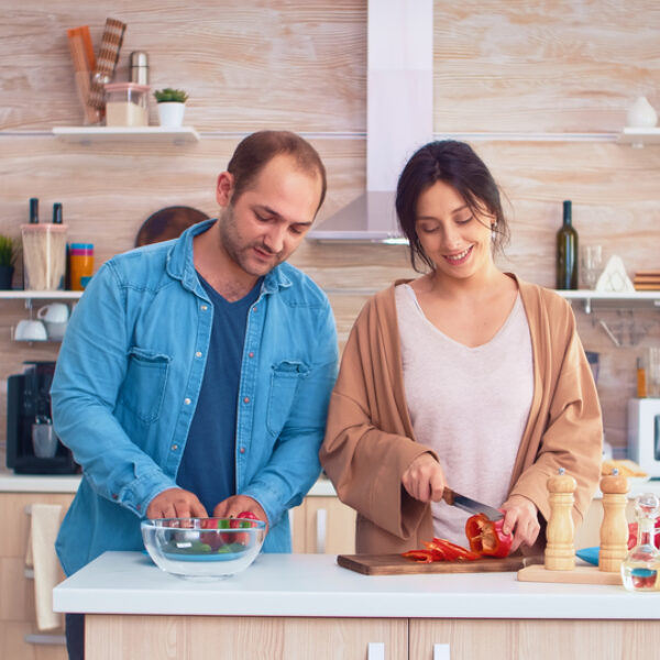 Wife chopping bell pepper on cutting board and husband opens refrigerator door. Cooking preparing healthy organic food happy together lifestyle. Cheerful meal in family with vegetables